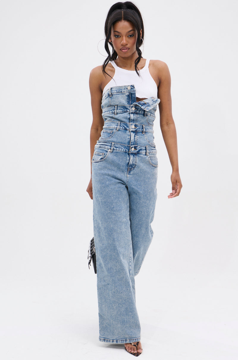 A woman confidently wears the DENIM DENIM DENIM LAYERED JUMPSUIT, featuring multiple overlapping denim waistbands and stacked buttons for a bold wide-leg look, styled with a white tank top and heeled sandals against a plain white background.