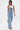 A woman confidently wears the DENIM DENIM DENIM LAYERED JUMPSUIT, featuring multiple overlapping denim waistbands and stacked buttons for a bold wide-leg look, styled with a white tank top and heeled sandals against a plain white background.