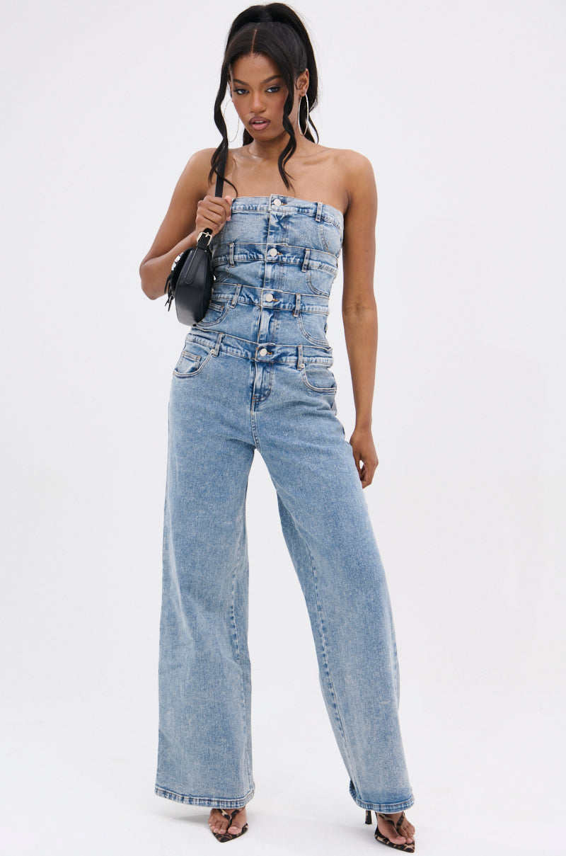 A woman poses against a white background wearing the DENIM DENIM DENIM LAYERED JUMPSUIT, a strapless wide-leg denim piece with button details. She accessorizes with a small black shoulder bag, long wavy hair, and open-toe heels.