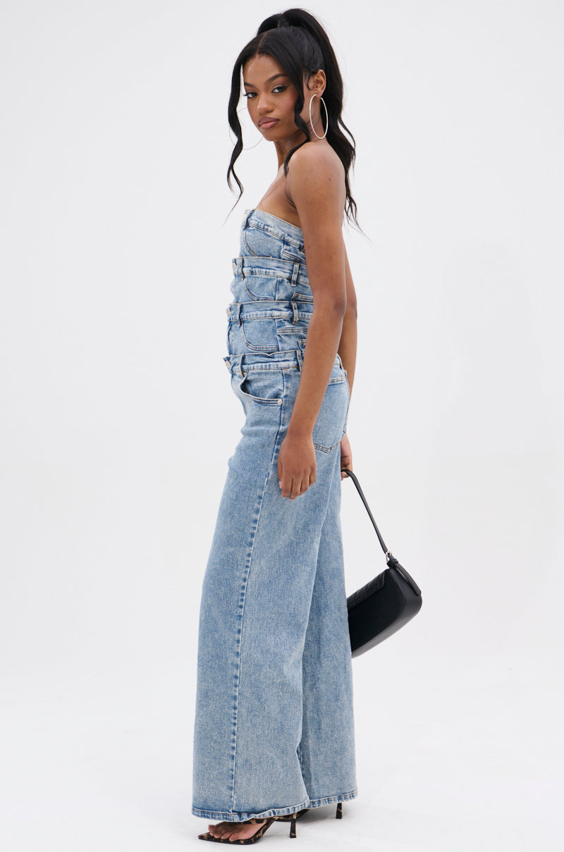 Woman stands sideways in the DENIM DENIM DENIM LAYERED JUMPSUIT, featuring a strapless top and wide-leg silhouette. She pairs it with black heels, hoop earrings, and a small black handbag against a plain white background.