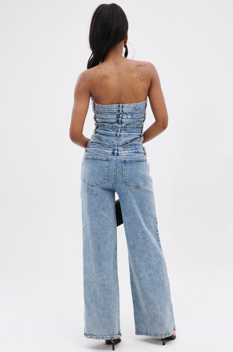 A woman faces away, wearing the DENIM DENIM DENIM LAYERED JUMPSUIT featuring a strapless, ruched denim bodice with back button details and high-waisted, wide-leg light-wash jeans against a plain white background.