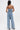 A woman faces away, wearing the DENIM DENIM DENIM LAYERED JUMPSUIT featuring a strapless, ruched denim bodice with back button details and high-waisted, wide-leg light-wash jeans against a plain white background.