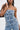 A woman wears the DENIM DENIM DENIM LAYERED JUMPSUIT with multiple waistbands, belt loops, and buttons for a stacked effect. She holds a black shoulder bag against a plain light background.