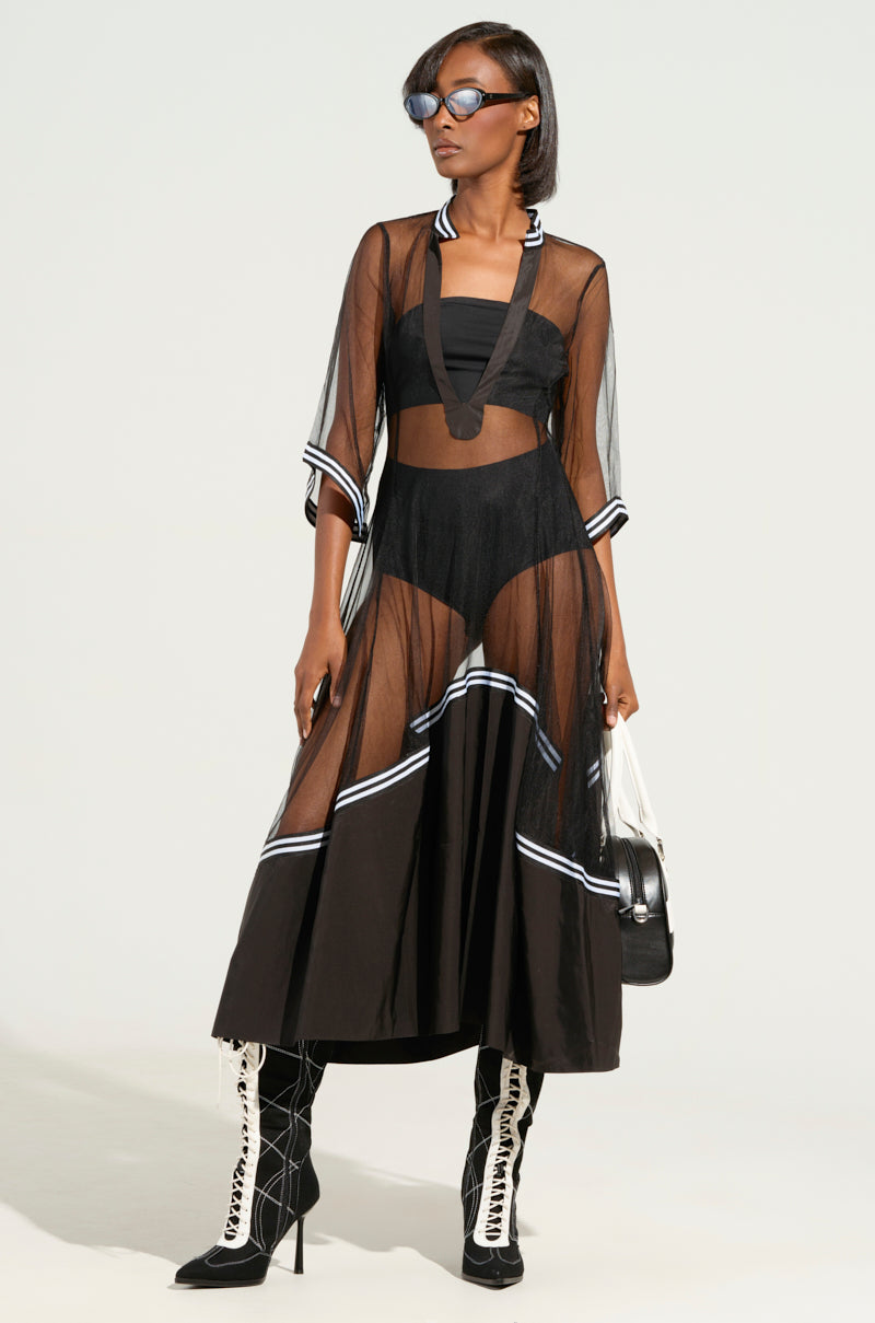 A person wearing the "NOW YOU SEE ME NOW YOU DON'T MIDI DRESS," characterized by its sheer fabric and white trim, layered over a black outfit. They have styled it with knee-high lace-up boots and sunglasses. Featuring a plunging neckline, they confidently pose against a light background while holding a handbag.