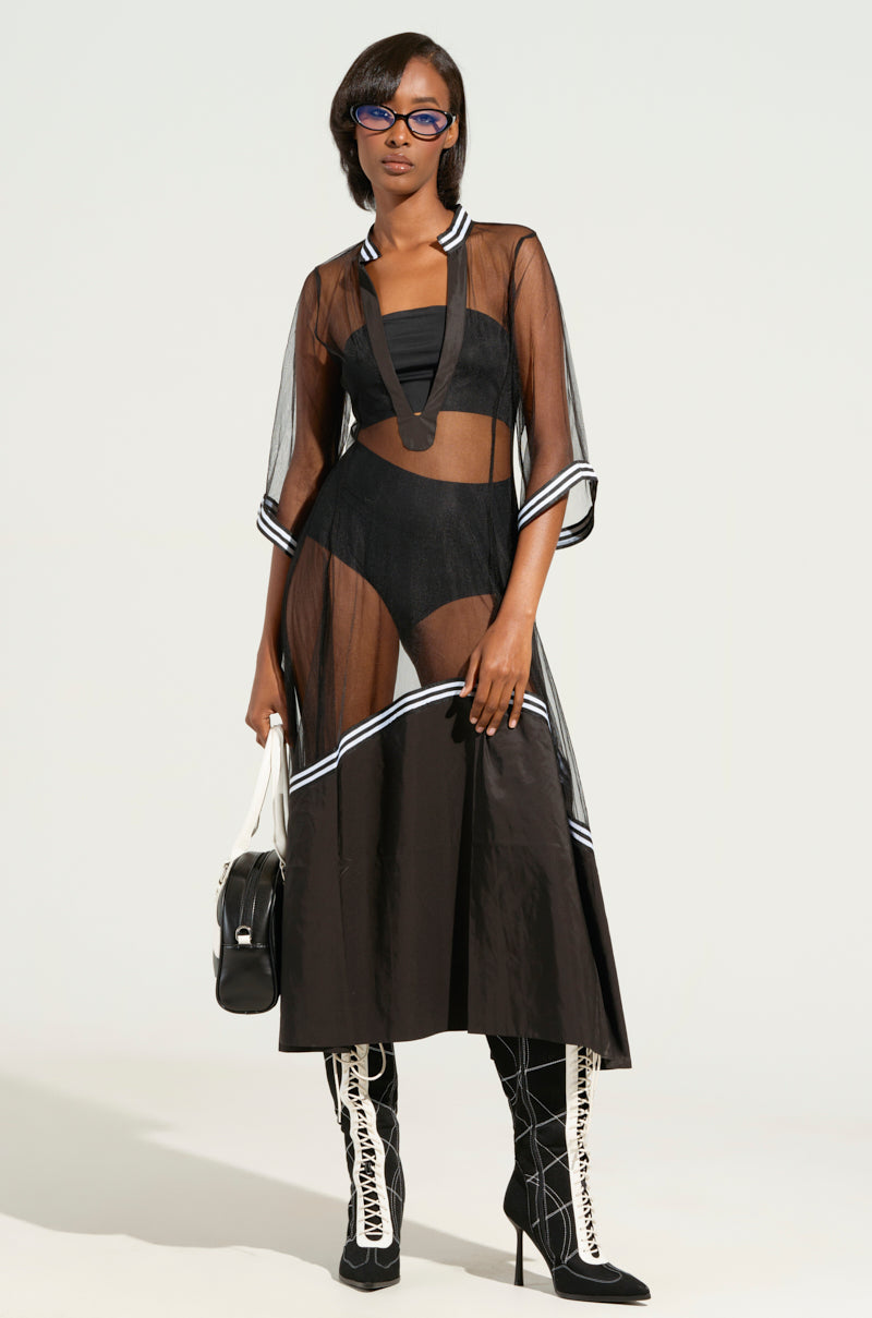A person stylishly dressed in the "NOW YOU SEE ME NOW YOU DON'T" midi dress, featuring a sheer design with a plunging neckline and white trim. They accessorize with a handbag, completing their look with tall black and white boots and sunglasses, all set against a simple background.