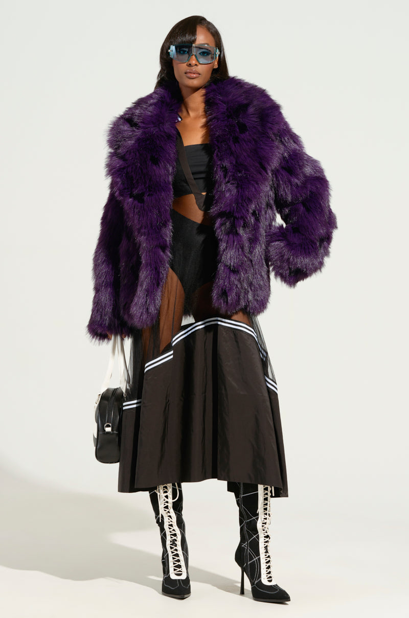 A person styled in the "NOW YOU SEE ME NOW YOU DON'T MIDI DRESS" is wearing a large purple fur coat over it. Completing the look are knee-high black and white lace-up boots, chic sunglasses, and a black handbag.
