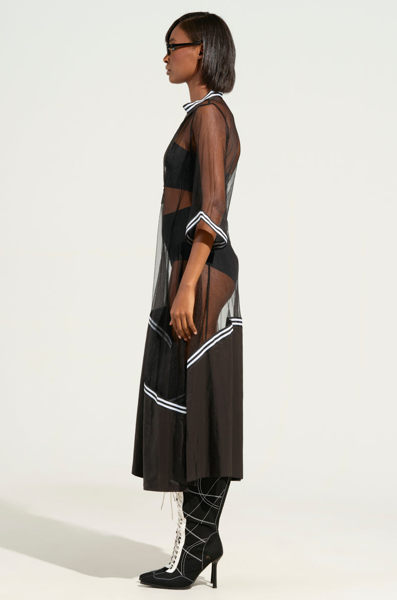 A person in side profile, elegantly dressed in the "NOW YOU SEE ME NOW YOU DON'T MIDI DRESS," with striped ribbon detailing over a black outfit, accessorizes with sunglasses and knee-high lace-up boots showcasing geometric patterns, standing against a plain background.