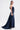 A woman stands in profile against a white background, wearing the LUNA DENIM MAXI DRESS with a strapless neckline and long train. She holds a small clutch; her straight brown hair and earrings complete the look.