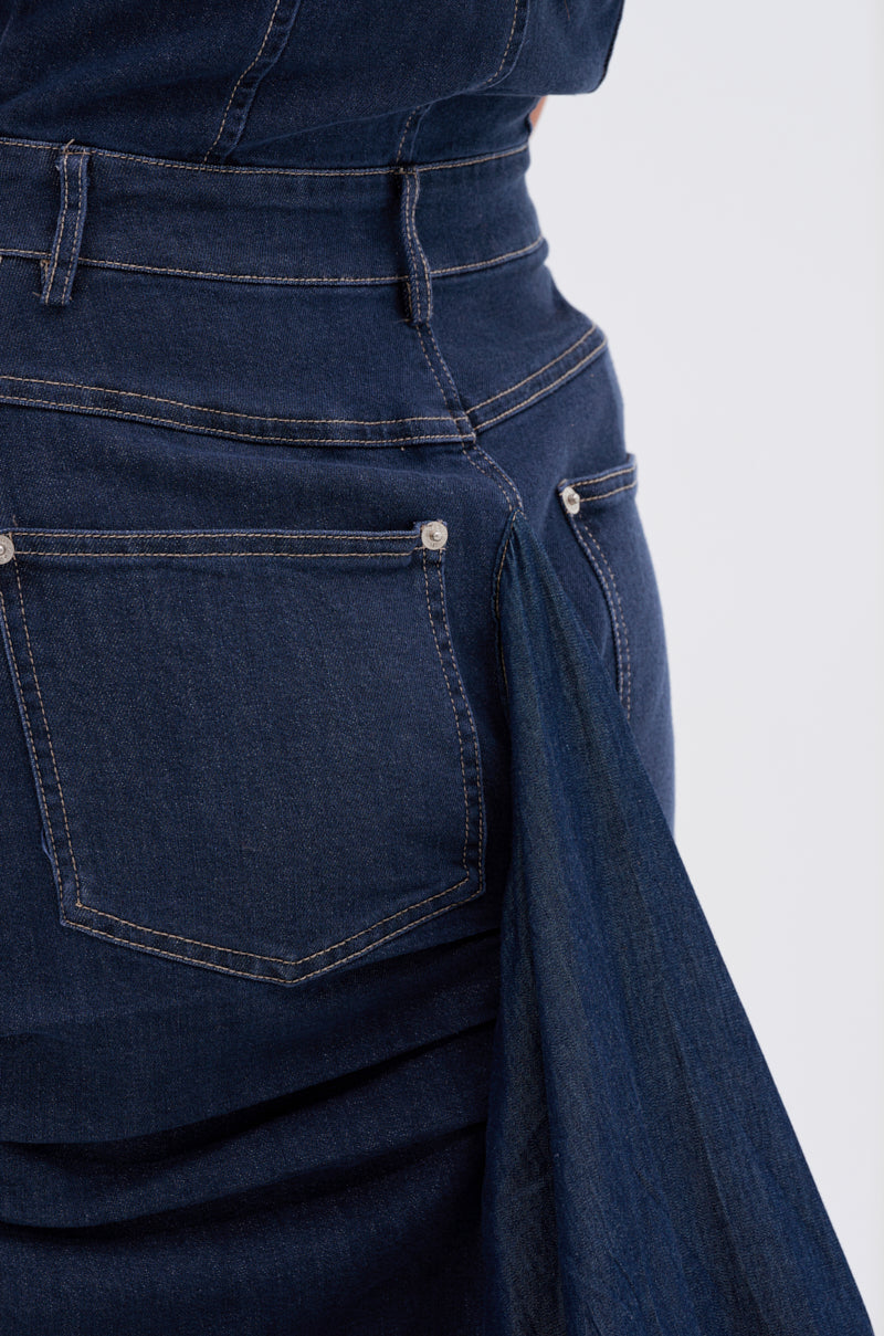 Close-up of the back of a person wearing the LUNA DENIM MAXI DRESS in dark blue denim with visible stitching. A long piece of denim fabric, resembling an A-line silhouette, is attached near the side pocket and drapes downward.
