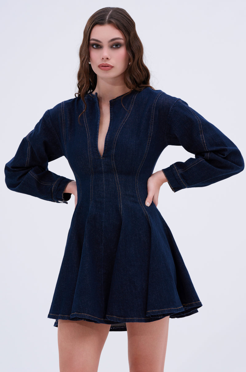 Confidently posing in the ALL ABOUT MY BUSINESS DENIM MINI DRESS IN DARK BLUE, this outfit boasts long sleeves, a flared skater silhouette, seam details, and a small V-neckline set against a plain white backdrop.