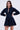 Confidently posing in the ALL ABOUT MY BUSINESS DENIM MINI DRESS IN DARK BLUE, this outfit boasts long sleeves, a flared skater silhouette, seam details, and a small V-neckline set against a plain white backdrop.