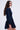 A woman with long, wavy hair wears the "ALL ABOUT MY BUSINESS DENIM MINI DRESS IN DARK BLUE," featuring long sleeves and a flared skater skirt. She stands in profile against a plain white background.