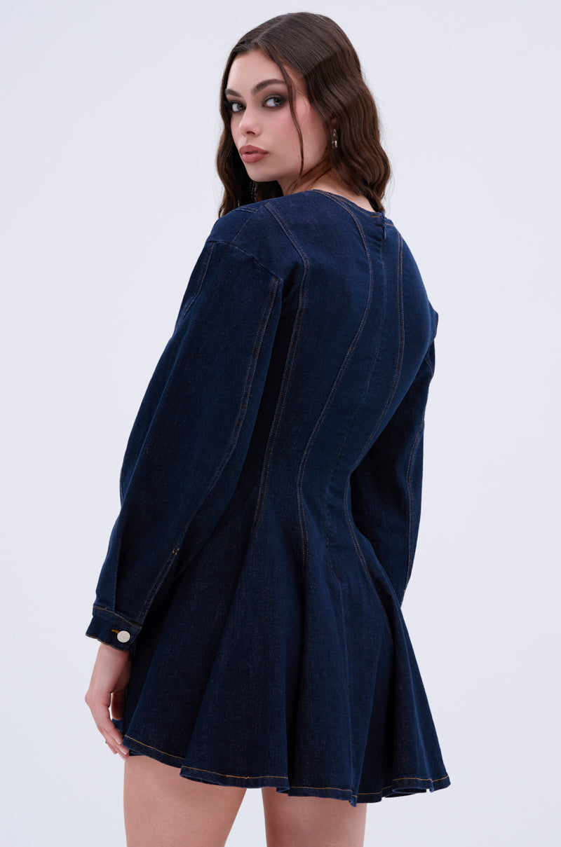 A woman with long brown hair wears the "All About My Business Denim Mini Dress in Dark Blue," featuring a skater silhouette with long sleeves and a flared skirt, against a light gray background, glancing over her shoulder.