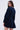 A woman with long brown hair wears the "All About My Business Denim Mini Dress in Dark Blue," featuring a skater silhouette with long sleeves and a flared skirt, against a light gray background, glancing over her shoulder.