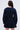 A person with long brown hair is seen from behind, showcasing the ALL ABOUT MY BUSINESS DENIM MINI DRESS IN DARK BLUE. This dress features a skater silhouette with long sleeves, a flared skirt, visible stitching, and buttoned cuffs against a plain white background.