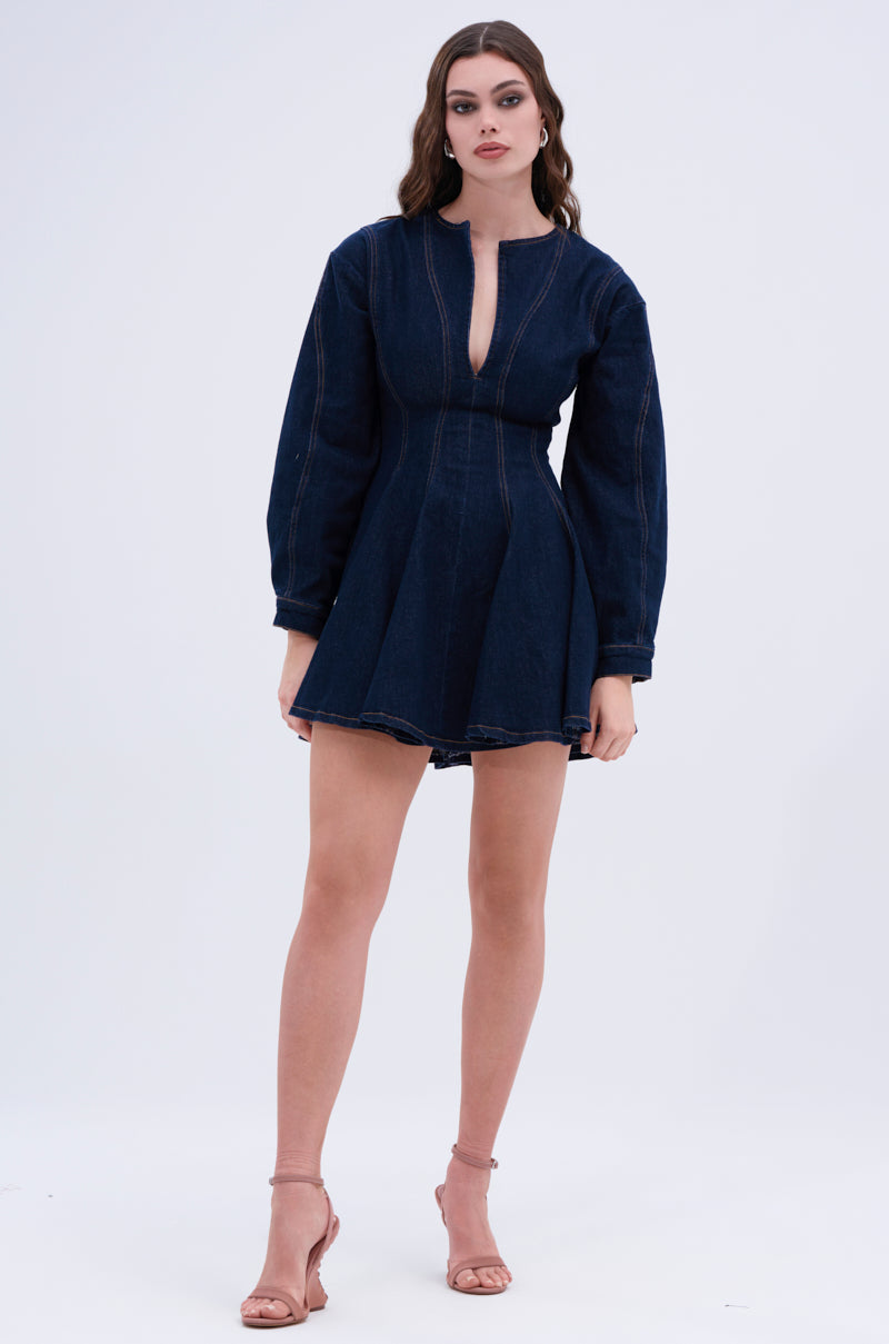 A woman poses against a plain backdrop wearing the ALL ABOUT MY BUSINESS DENIM MINI DRESS in dark blue, with long sleeves and a front cutout. Her long, wavy hair complements her beige high-heeled sandals, creating a chic skater look.