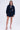 A woman poses against a plain backdrop wearing the ALL ABOUT MY BUSINESS DENIM MINI DRESS in dark blue, with long sleeves and a front cutout. Her long, wavy hair complements her beige high-heeled sandals, creating a chic skater look.