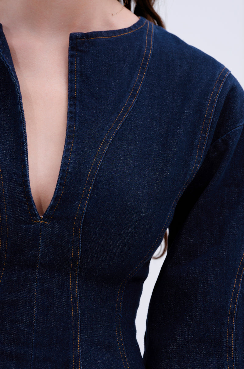 Close-up of a person in the ALL ABOUT MY BUSINESS DENIM MINI DRESS IN DARK BLUE, highlighting its structured fit, seam details, deep V-neck, and skater silhouette against a light gray background that emphasizes the dress's textured elegance.