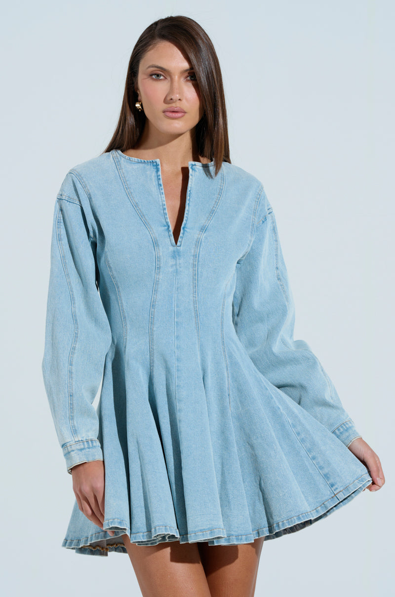 Someone wearing the ALL ABOUT MY BUSINESS DENIM MINI DRESS, featuring a light blue wash, long sleeves, a flared skirt, and a V-neckline. The person has long brown hair against a plain light gray background.
