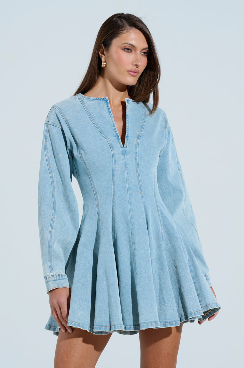 A person with long hair wears the "ALL ABOUT MY BUSINESS DENIM MINI DRESS," which has long sleeves, a v-neck, and a flared skirt. The light wash denim stands out against a simple background, highlighting the outfit.