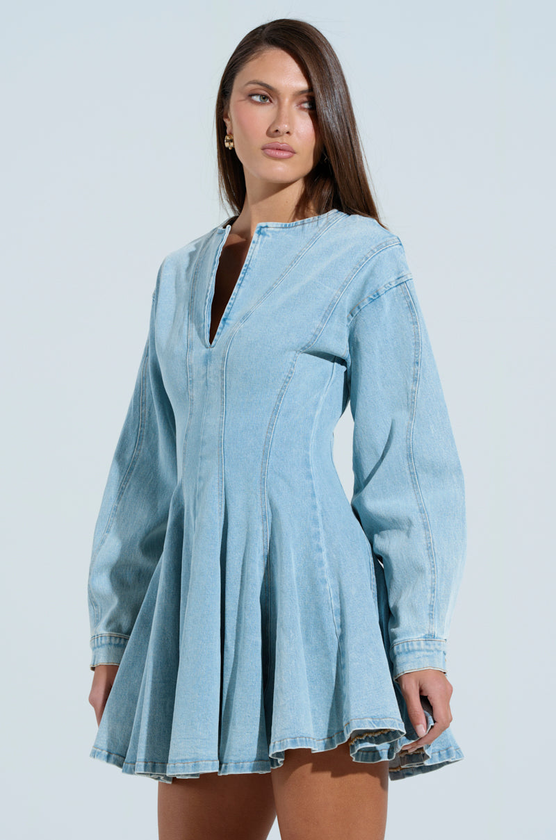 Wearing the "ALL ABOUT MY BUSINESS DENIM MINI DRESS" with a fitted waist and flared skirt, a person stands against a matching background. This light blue mini dress features a V-neck and long sleeves, beautifully framing their long brown hair as they gaze slightly to the side.