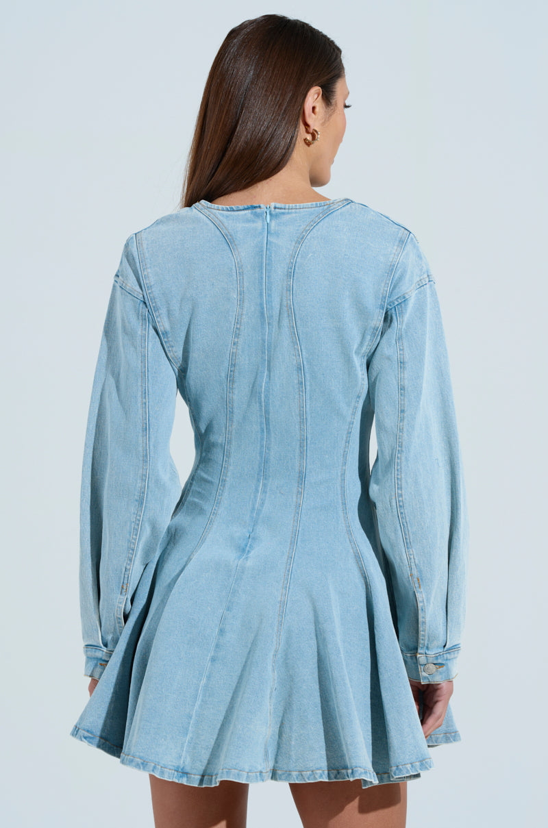 A woman in the "All About My Business" denim mini dress, featuring a fitted long-sleeve design, high neckline, flared skirt, and light wash color. Facing away to highlight the back details, her straight brown hair cascades over her shoulders.