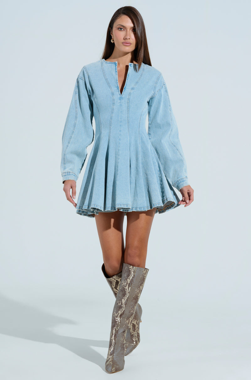A person confidently stands in the ALL ABOUT MY BUSINESS DENIM MINI DRESS, featuring a flared skirt and long sleeves. They pair it with knee-high snakeskin boots and a chic high neckline against a plain light blue background, complementing the stylish ensemble.