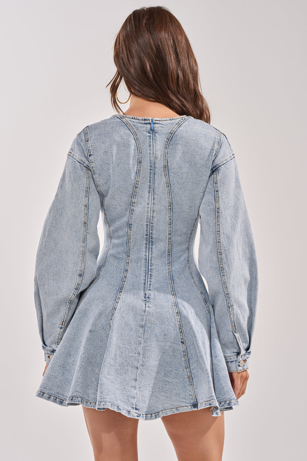 A woman stands with her back to the camera, wearing the ALL ABOUT MY BUSINESS DENIM MINI DRESS featuring a light wash, fitted waist, and flared skirt. Her long brown hair is down against a plain, light background. 5