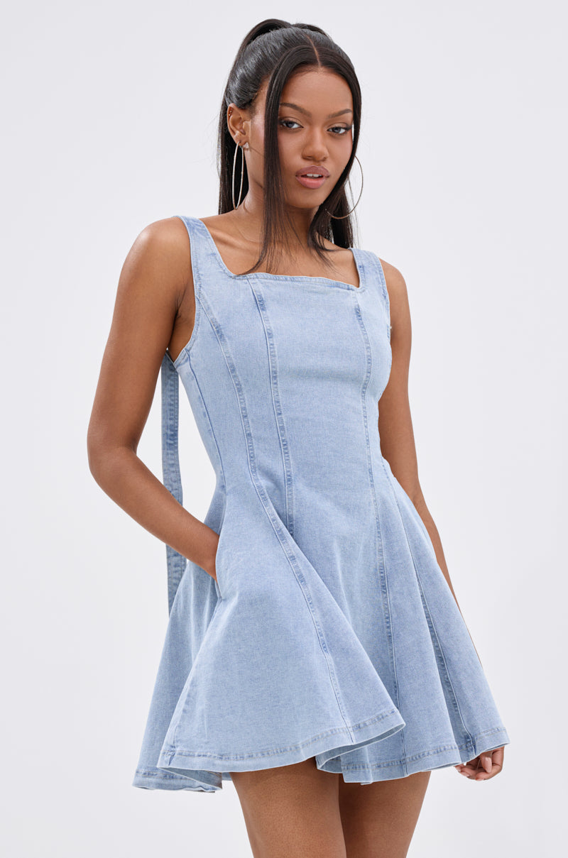 A woman with long dark hair in a high ponytail wears the JOANNA DENIM MINI DRESS, featuring a square neckline, flared skirt, and pockets. She stands against a plain light background with her hands tucked into the dress pockets. 0
