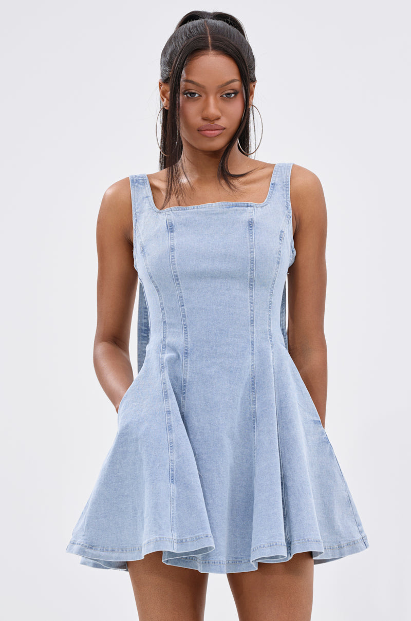 A woman stands against a plain light background with her hands in the pockets of the sleeveless JOANNA DENIM MINI DRESS, featuring a fit-and-flare silhouette, square neckline, and flared skirt. 2