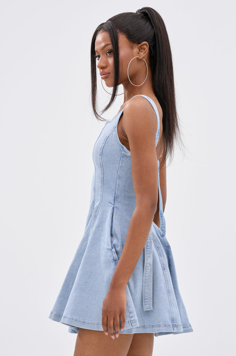 A woman poses sideways against a plain background in the JOANNA DENIM MINI DRESS, a sleeveless, light blue fit-and-flare mini dress with a square neckline. She wears large hoop earrings and has long hair styled in a high ponytail. 3