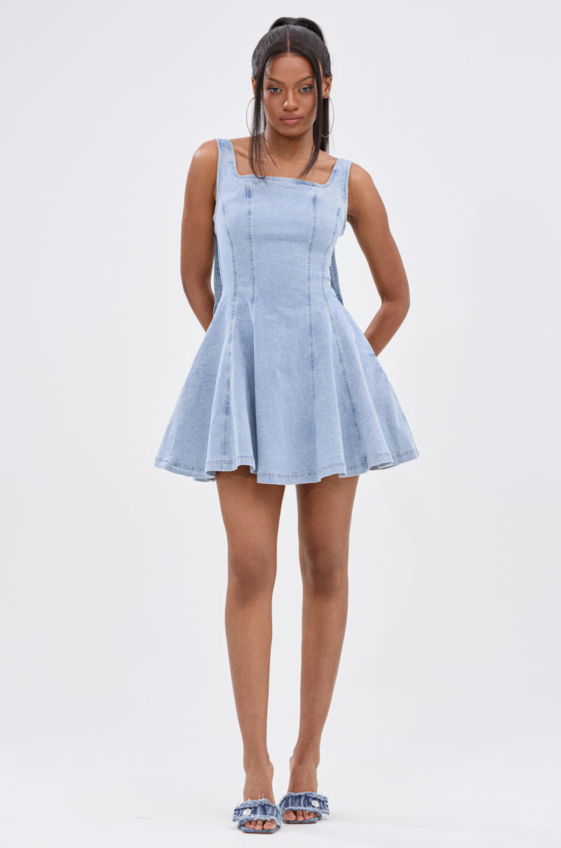 A woman stands against a white background wearing the JOANNA DENIM MINI DRESS with a square neckline and fit-and-flare silhouette. Her long hair is in a half-up ponytail, and she pairs the dress with blue sandals. 6
