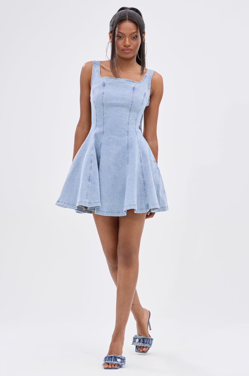 A woman with long, straight hair walks forward in the JOANNA DENIM MINI DRESS, a light blue fit-and-flare mini with a square neckline and flared skirt. She pairs it with blue heeled sandals featuring fringe details. The background is plain white. 7