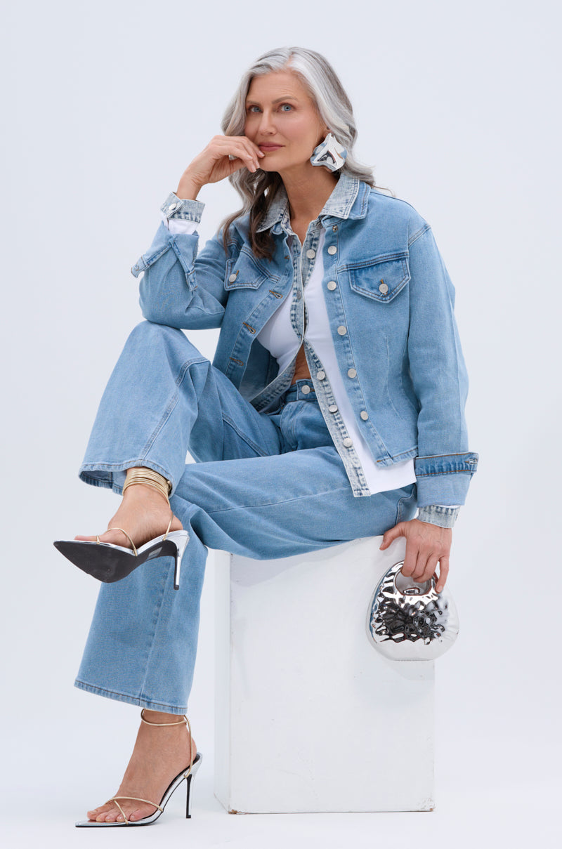 A stylish older woman with long gray hair wears TAKE ME TO THE RODEO JEANS with a light blue denim jacket, silver heels, statement earrings, and holds a shiny silver handbag while sitting on a white block.
