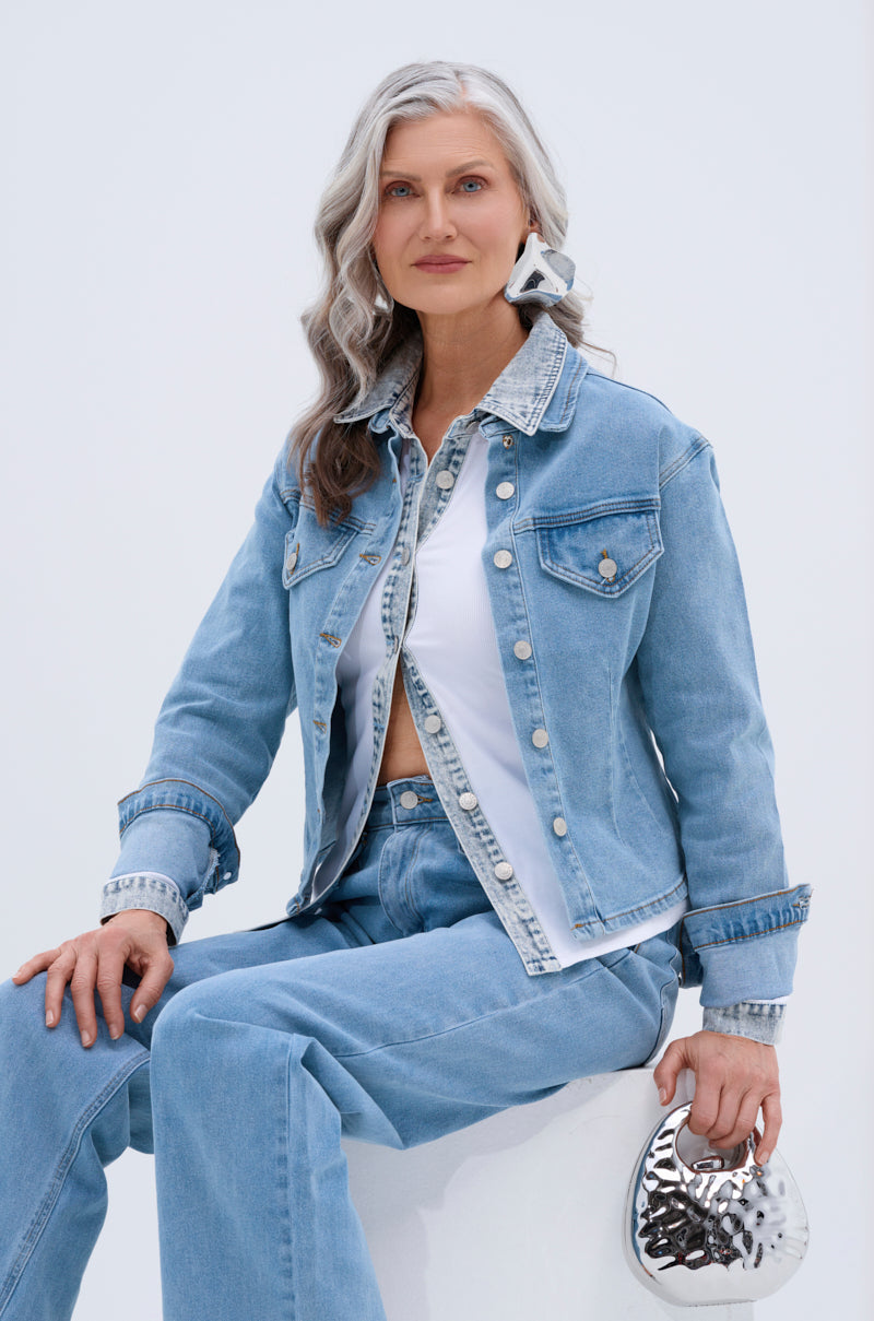 A woman with long gray hair and a slight smile wears the TAKE ME TO THE RODEO BUTTON UP denim jacket over a white shirt with jeans and large earrings, sitting and holding a small silver clutch against a plain light background.