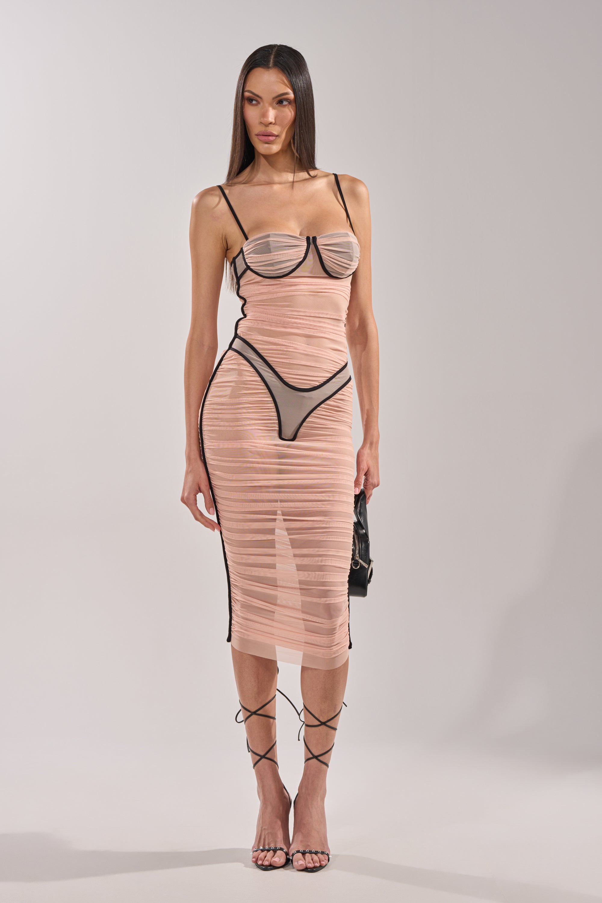 A woman poses against a plain background in the BIKINI BABE MESH MIDI DRESS, featuring sheer mesh fabric, black straps, visible bustier details, straight long hair, strappy heels, and a small black purse. 0