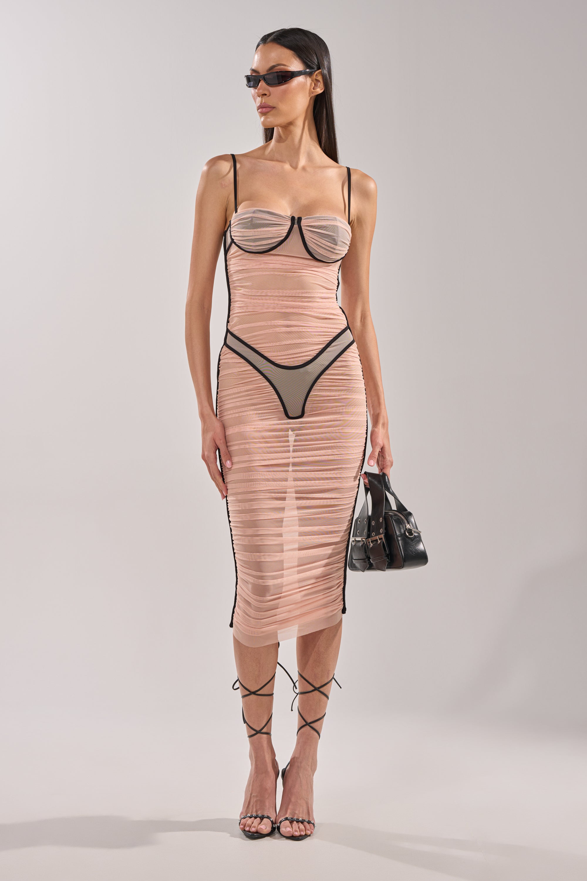 A woman stands against a plain background wearing the BIKINI BABE MESH MIDI DRESS with black trim, paired with black high-waisted underwear, strappy sandals, sunglasses, and a small black handbag. 1
