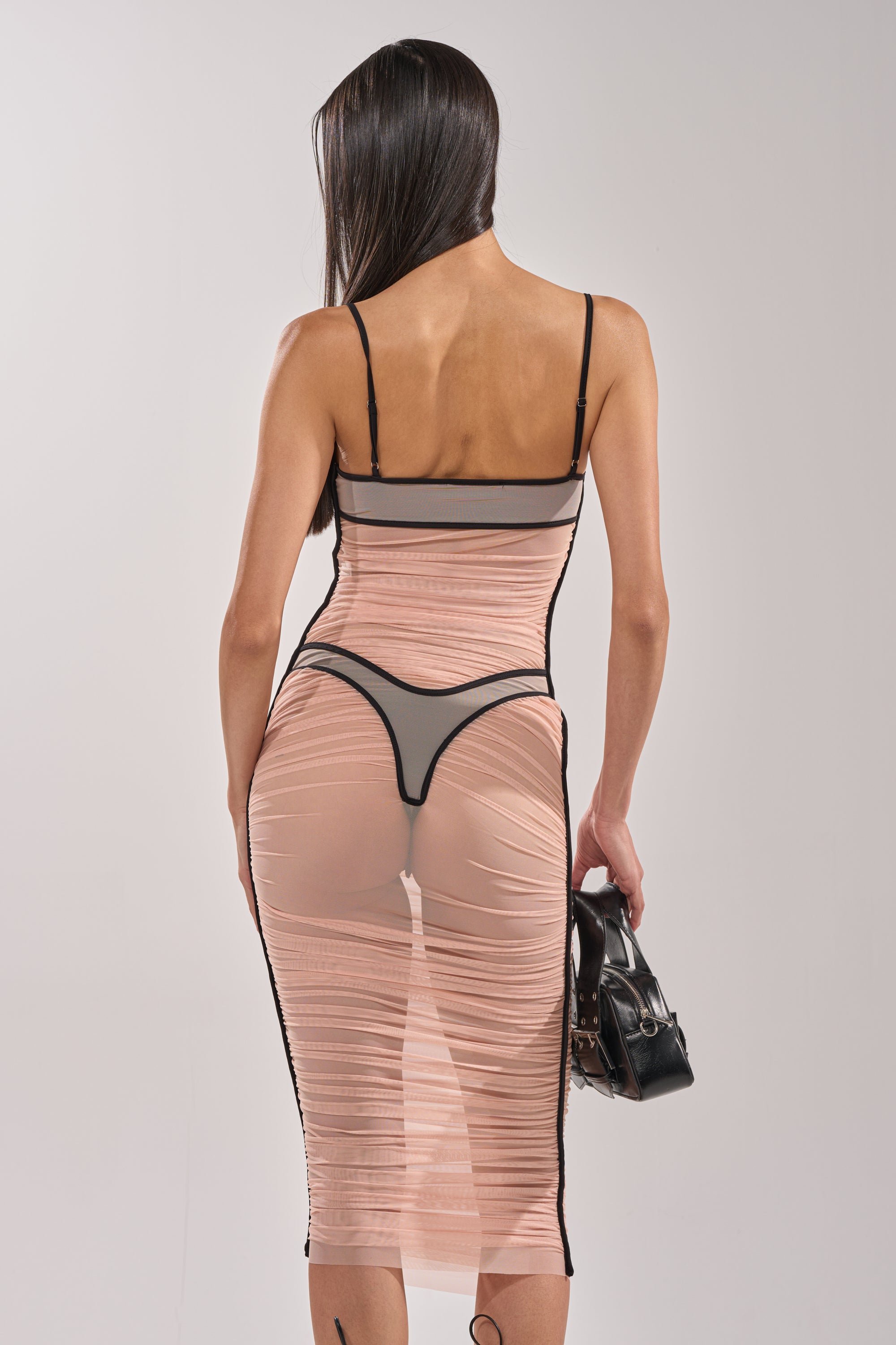 A woman with long dark hair faces away, wearing the BIKINI BABE MESH MIDI DRESS—a curve-hugging, ruched midi in sheer nude mesh with black trim and visible black thong—holding a small black handbag against a plain background. 6
