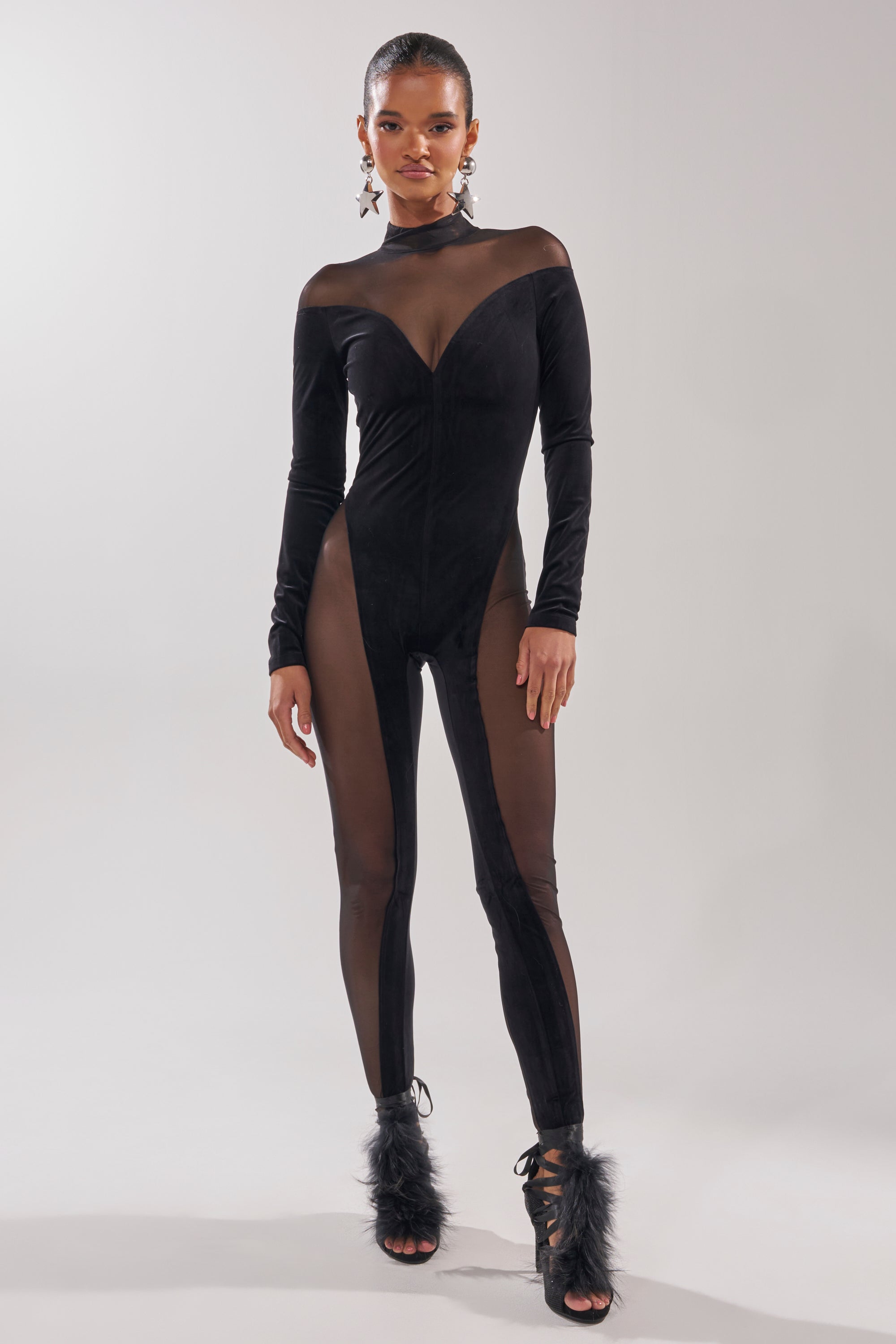 A woman poses in the MESH TO IMPRESS LONG SLEEVE JUMPSUIT, a black sheer piece with paneled mesh, long sleeves, and a high neckline. She accessorizes with star earrings and black feather-accented heels against a light background. 2