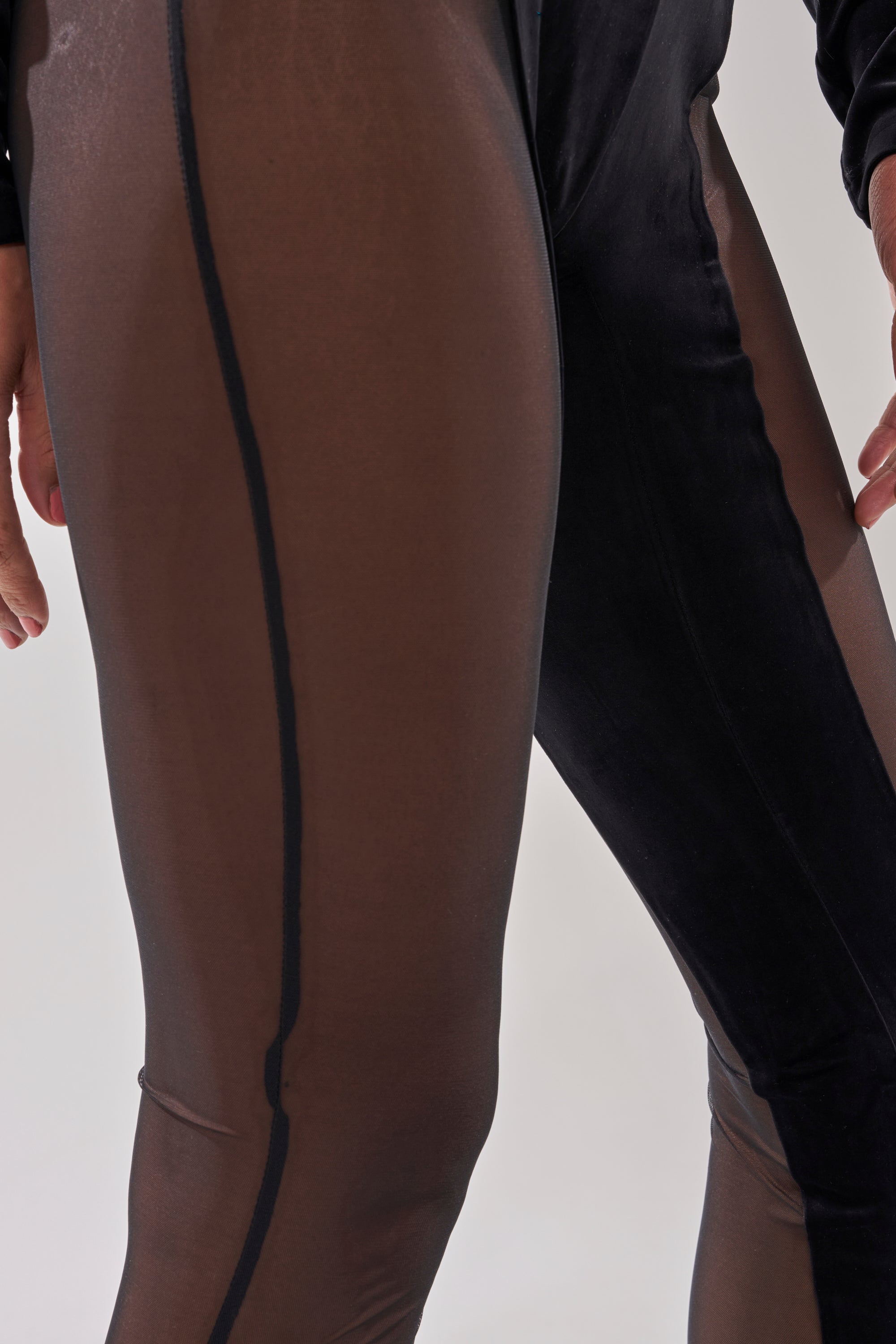 Two people stand side by side against a light background, wearing the MESH TO IMPRESS LONG SLEEVE JUMPSUIT in sheer brown and black with vertical seams. Only their legs and hands are visible. 6
