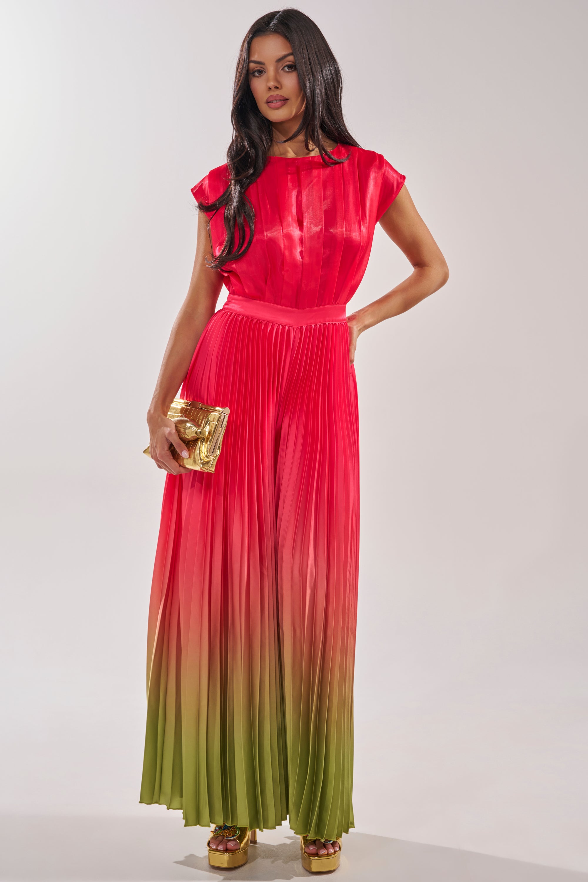 A woman in the SUNSET VIBES SATIN JUMPSUIT, with long dark hair, gold clutch, and platform shoes, is perfectly styled for a summer wedding as she poses against a plain light background. 1