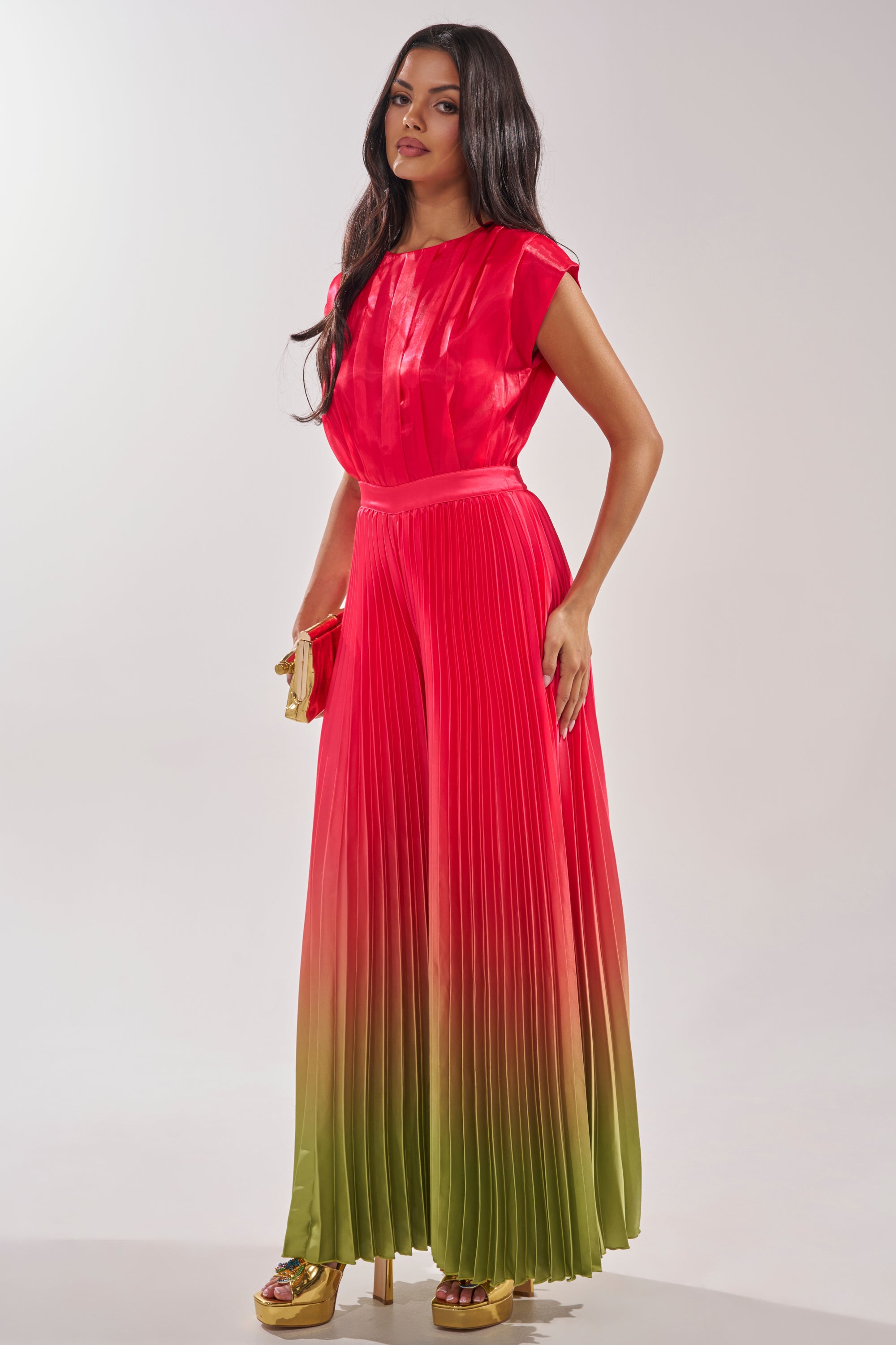 A woman wears the SUNSET VIBES SATIN JUMPSUIT, featuring a pleated silhouette fading from bright red to olive green, styled with a gold clutch and gold platform heels. 2