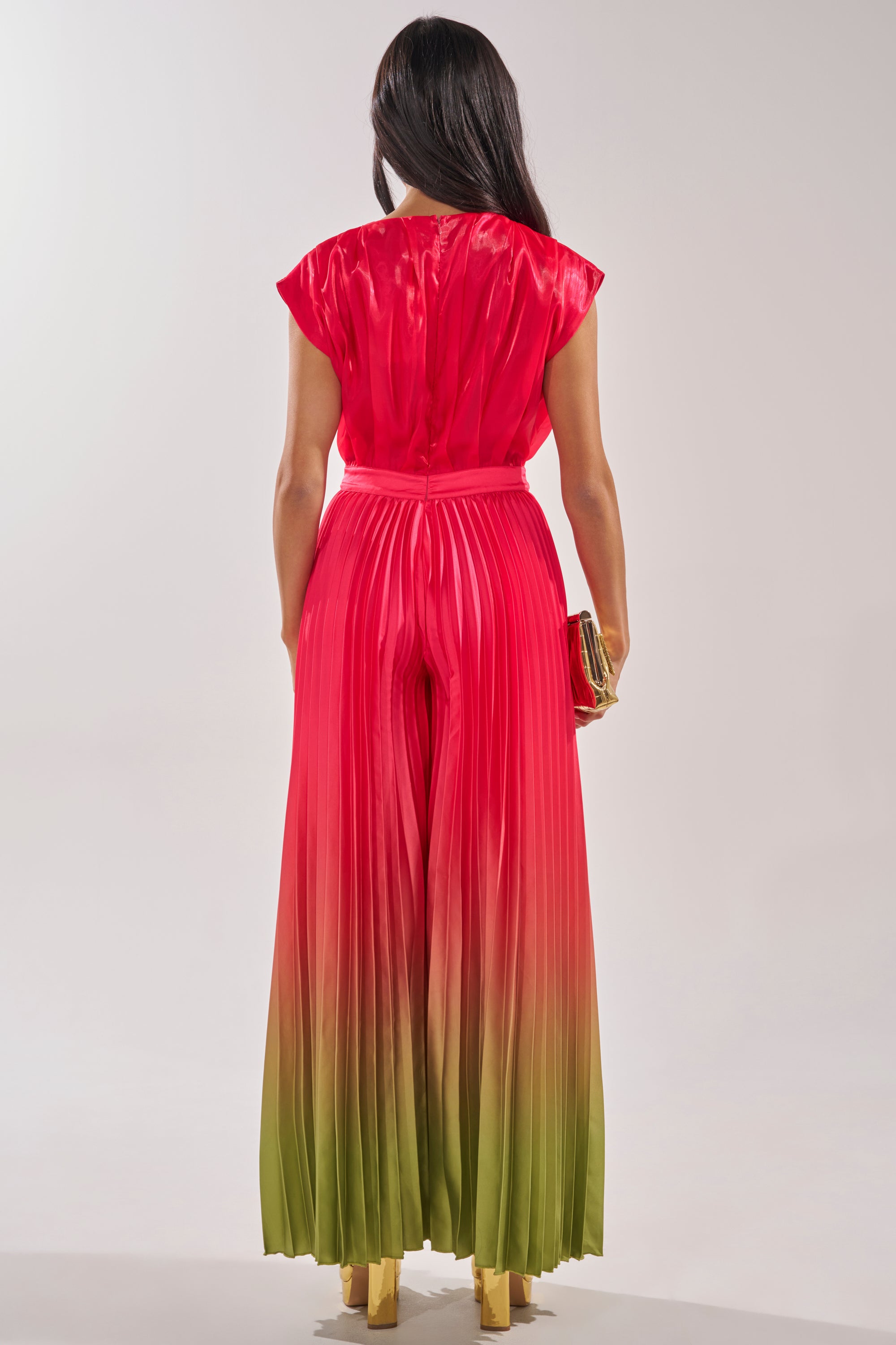 A woman with flowing dark hair faces away, wearing the SUNSET VIBES SATIN JUMPSUIT—a short sleeve, wide leg ombre jumpsuit fading from pink to green—styled with a clutch. The ideal choice for a summer wedding guest look. 3