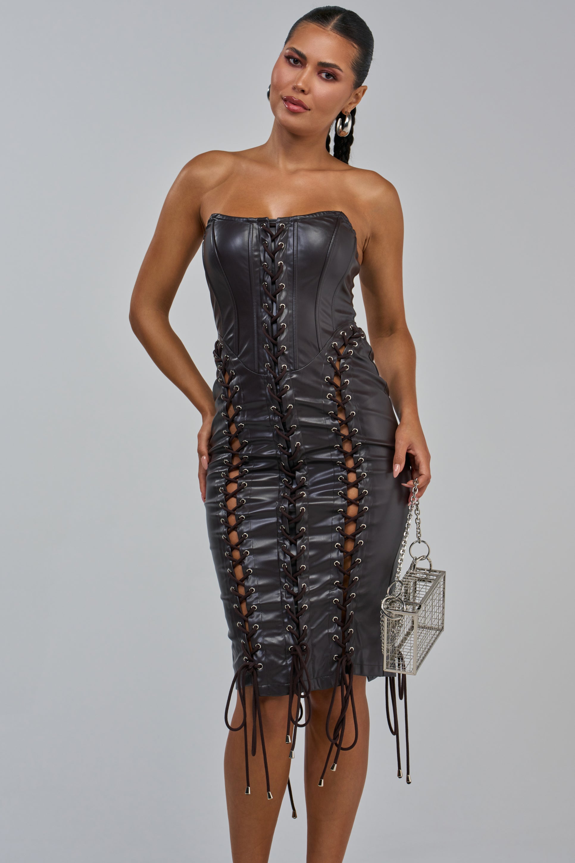 A woman wearing the SIN CITY Lace Up PU Midi Dress in brown—a strapless, fitted faux leather dress with lace-up details down the front and sides—holds a small, silver box-shaped handbag as she stands against a plain gray background.