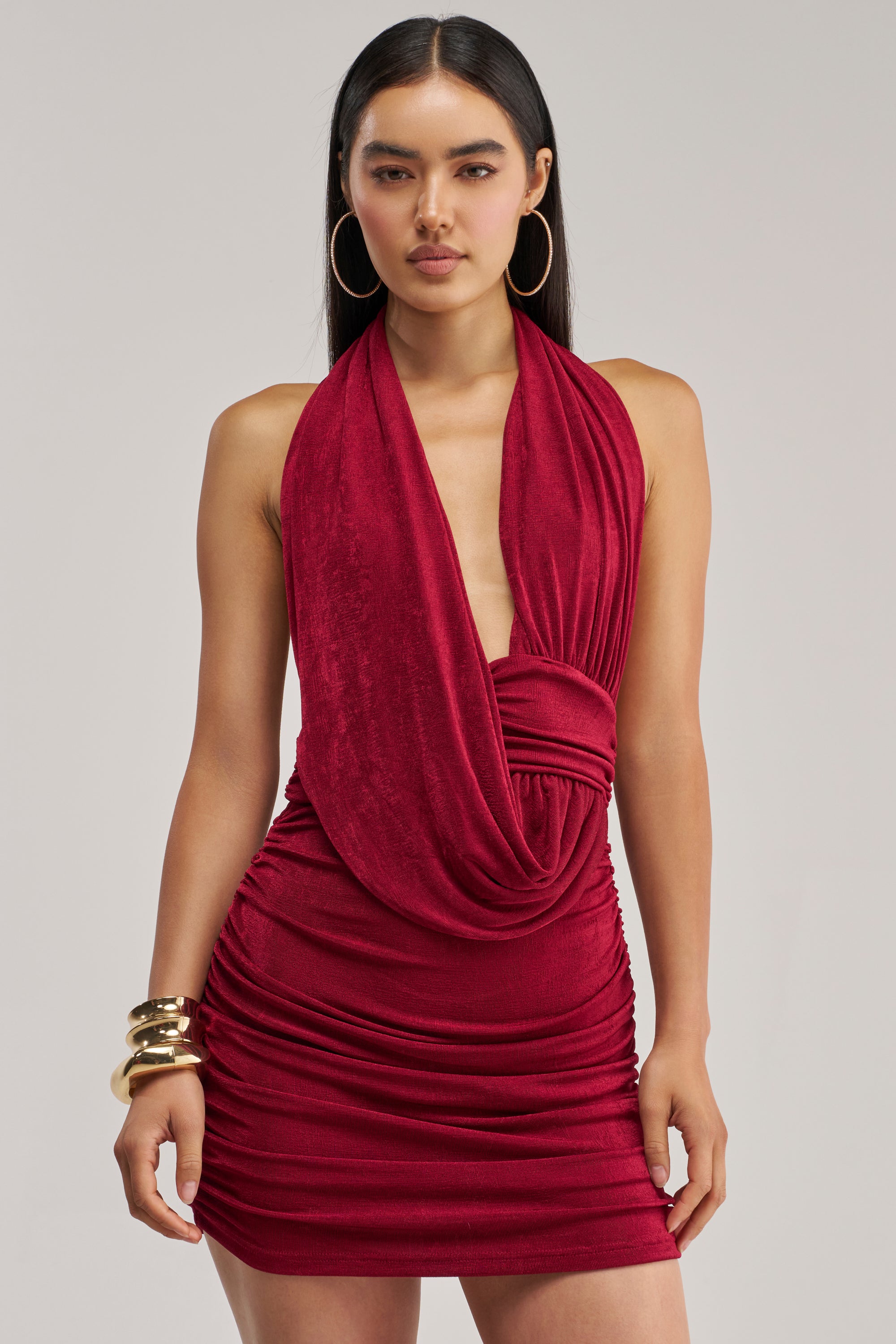 A woman wearing the HAUTE HEAT SWOOPED MINI DRESS IN BURGUNDY—a sleeveless, sexy halter mini dress with a draped neckline—paired with gold hoop earrings and chunky gold bangles, standing against a plain background.