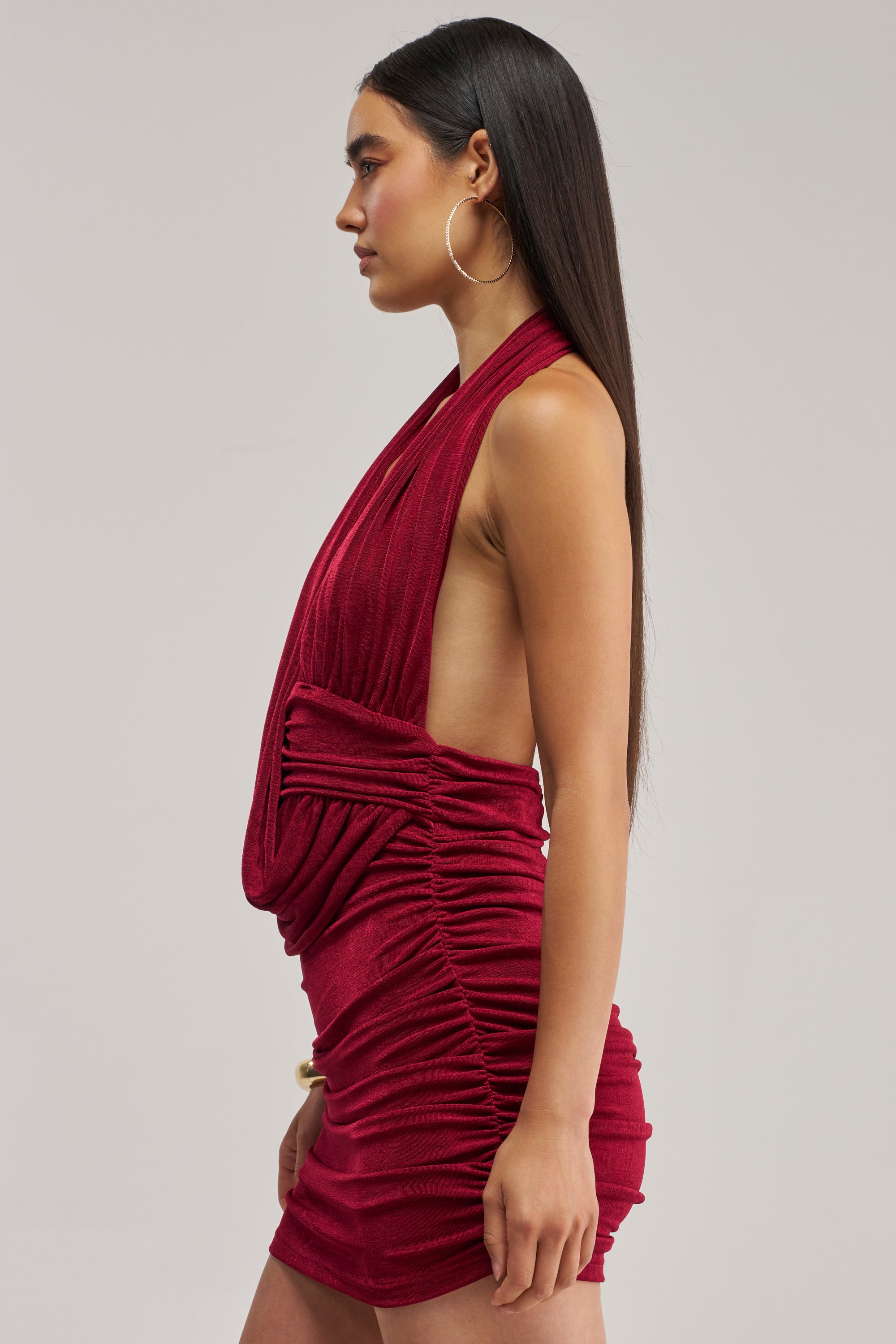 A woman stands in profile wearing the HAUTE HEAT SWOOPED MINI DRESS IN BURGUNDY—a sleeveless, backless mini dress with ruched detailing that perfectly accentuates her figure. She completes the look with large hoop earrings, her straight, long dark hair cascading down her back, and neutral makeup that highlights her features. The photograph is set against a plain light background.