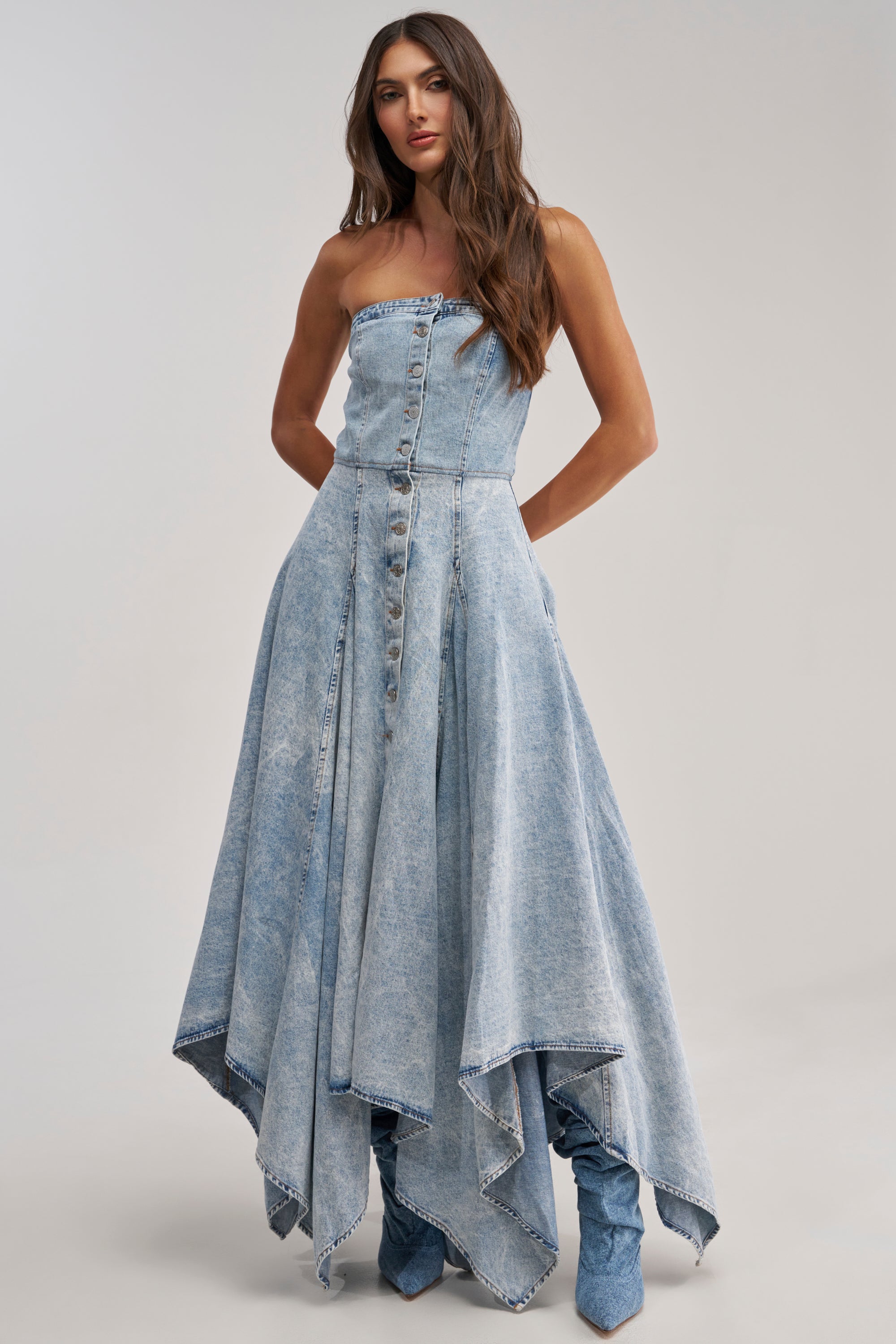 A woman models the MISS ME YET STRAPLESS DENIM MIDI DRESS with a handkerchief hem and silver button front, styled with matching denim boots against a plain light backdrop.