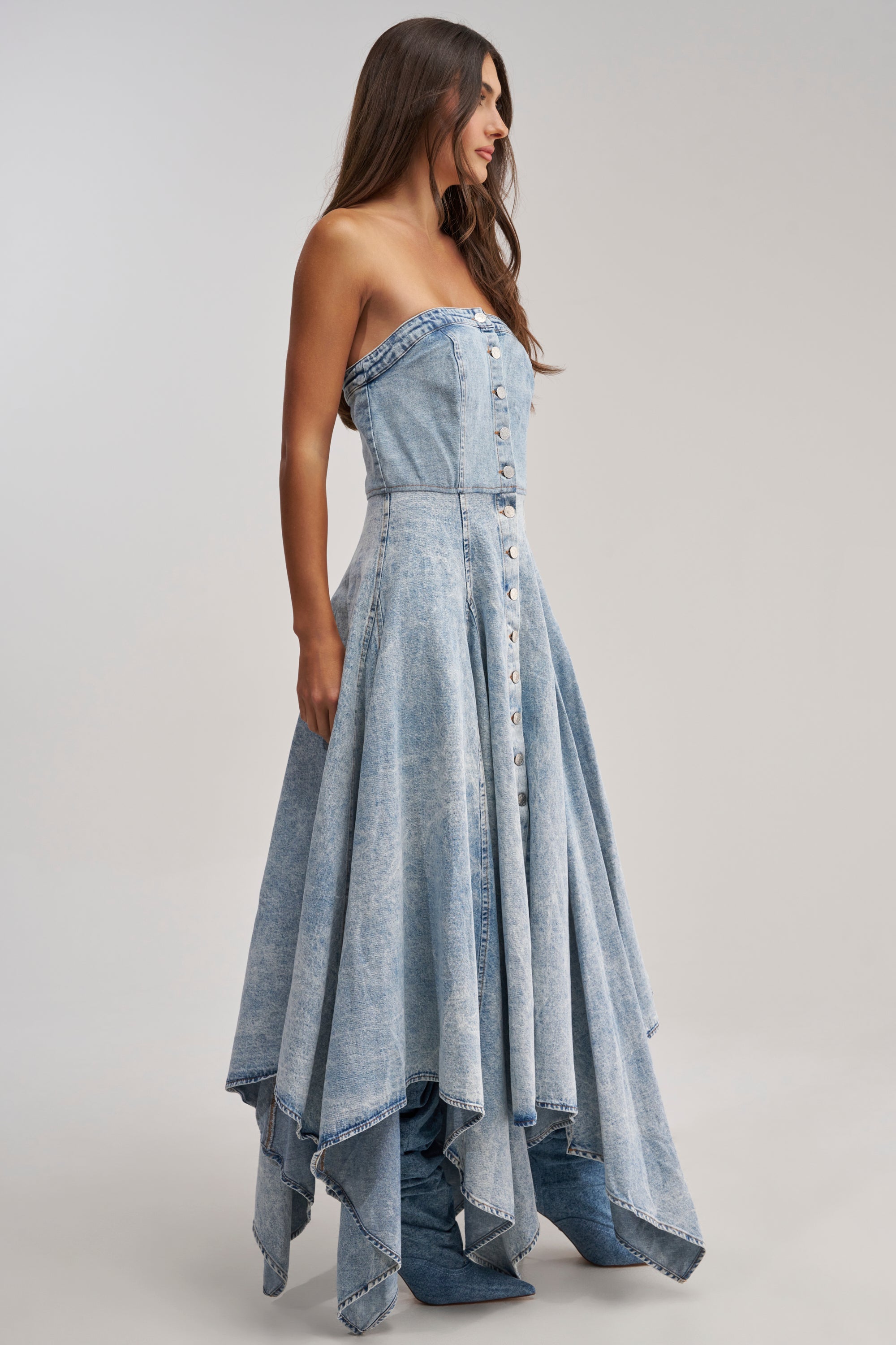 A woman with long brown hair models the MISS ME YET STRAPLESS DENIM MIDI DRESS, featuring silver front buttons and an asymmetrical handkerchief hemline, against a plain light background.