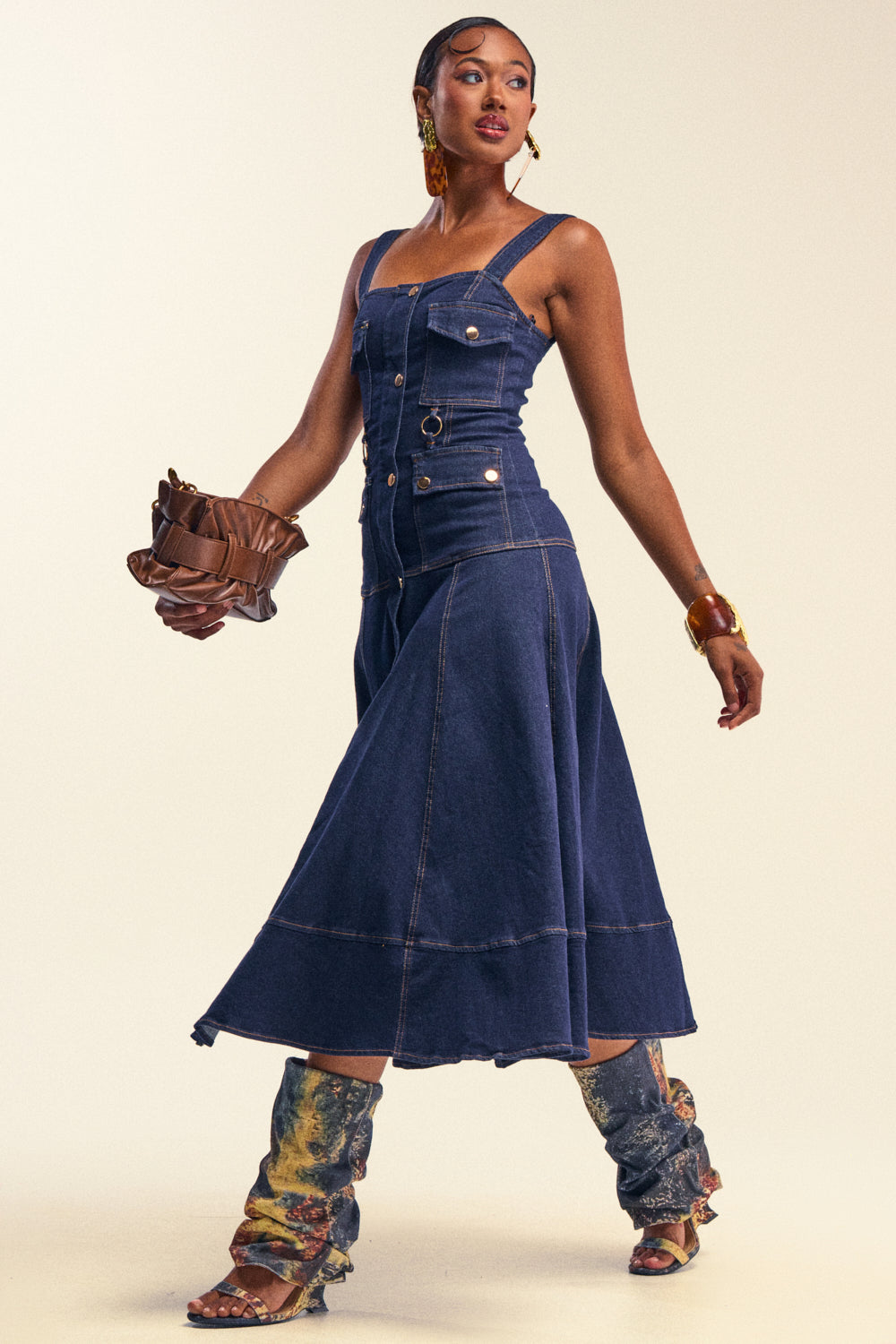 A woman models the WILDFLOWER DRIFT MIDI DRESS IN DARK BLUE DENIM—a sleeveless, buttoned midi dress with a subtle mermaid silhouette and flared skirt—styled with printed boots, statement earrings, and a brown clutch. She poses confidently against a neutral backdrop for effortlessly chic summer style.