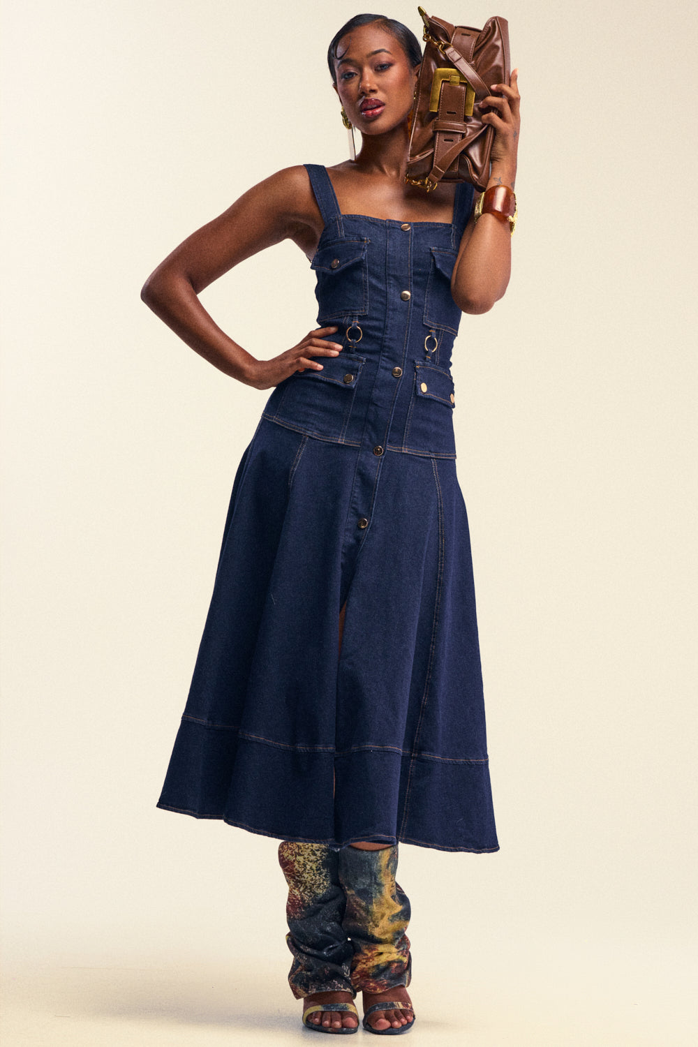 Woman models the WILDFLOWER DRIFT MIDI DRESS IN DARK BLUE DENIM, styled sleeveless and utility-inspired, holding a brown clutch bag to her head, accessorized with a gold bracelet and distinctive patterned boots.